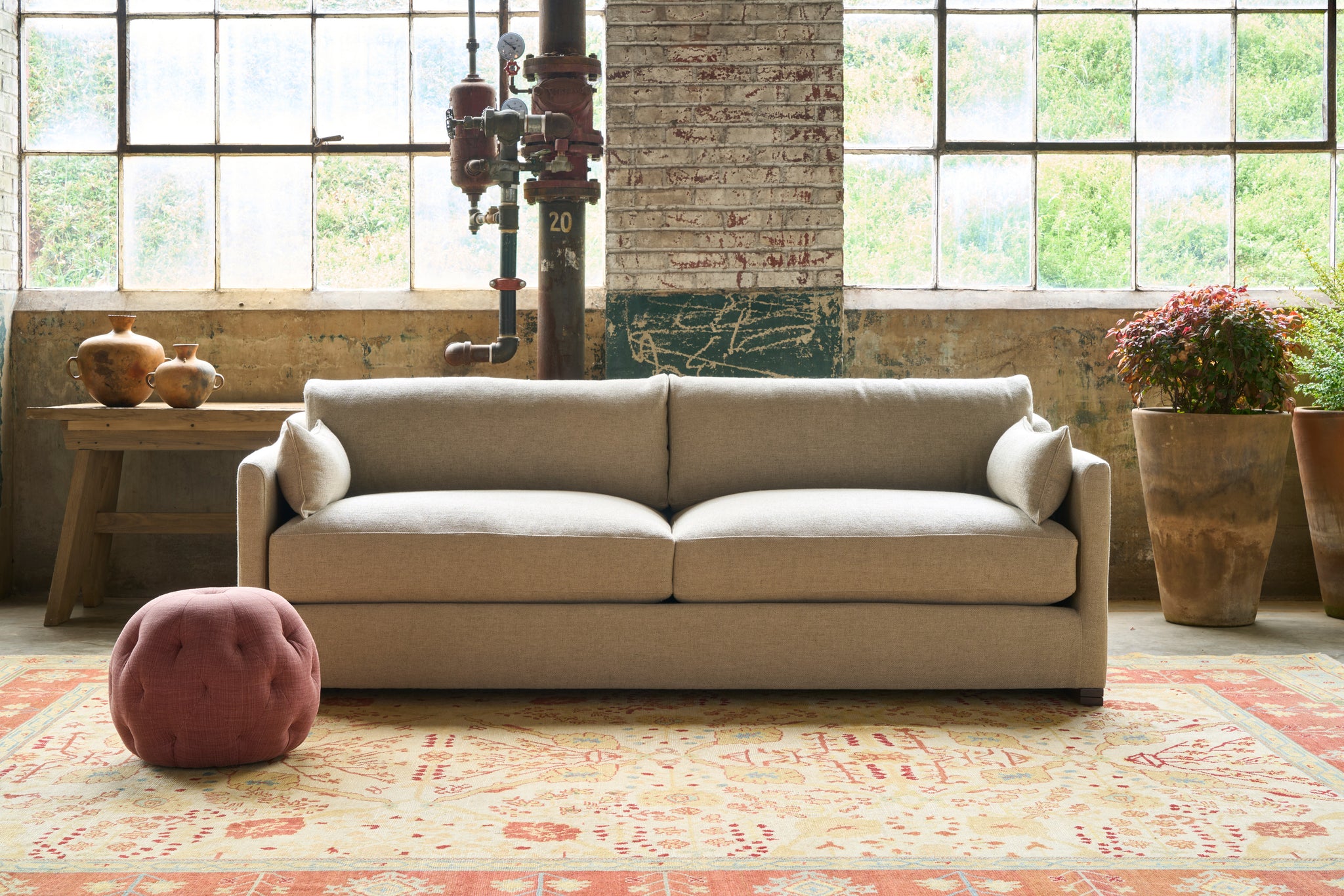  Oatmeal sofa in front of 2 windows with a red pouf in front. Wood table on the left with 2 pots on top. Photographed in Bellamy Oatmeal. 