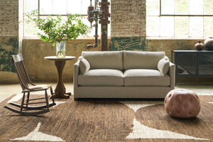  Oatmeal colored loveseat in a showroom with a wood rocking chair on the left and a pink  leather pouf on the right. Photographed in Solvang Mauve. 