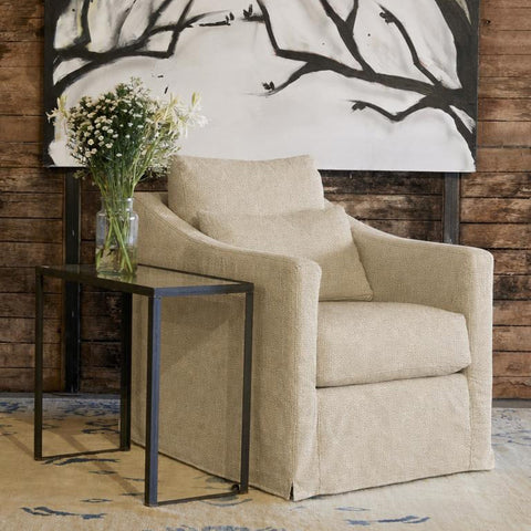 A beige upholstered armchair with a matching cushion, styled next to a black metal side table holding a vase of white flowers, set against a rustic wood plank wall with a large black-and-white tree branch painting.