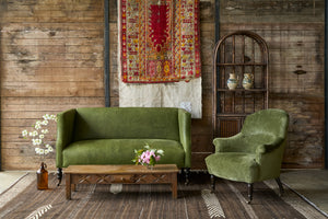  Green leather loveseat with a green velvet chair in front of a wood wall with some rug art. Wood coffee table with a drawer in front. Rattan cabinet on the right. Photographed in Archer Forest. 