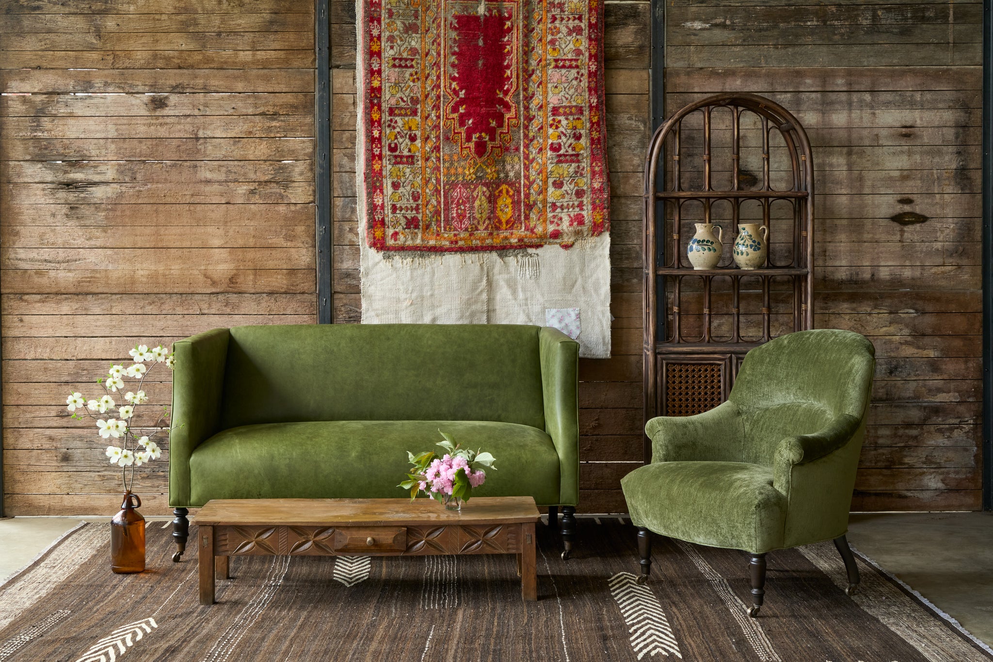  Green leather loveseat with a green velvet chair in front of a wood wall with some rug art. Wood coffee table with a drawer in front. Rattan cabinet on the right. Photographed in Velluto Olive. 