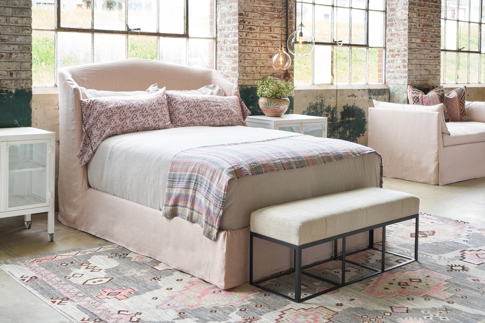  Slipcovered bed in a showroom with large windows behind. A white bench is at the end and a pink daybed is in the background. Photographed in Brevard Rose. 