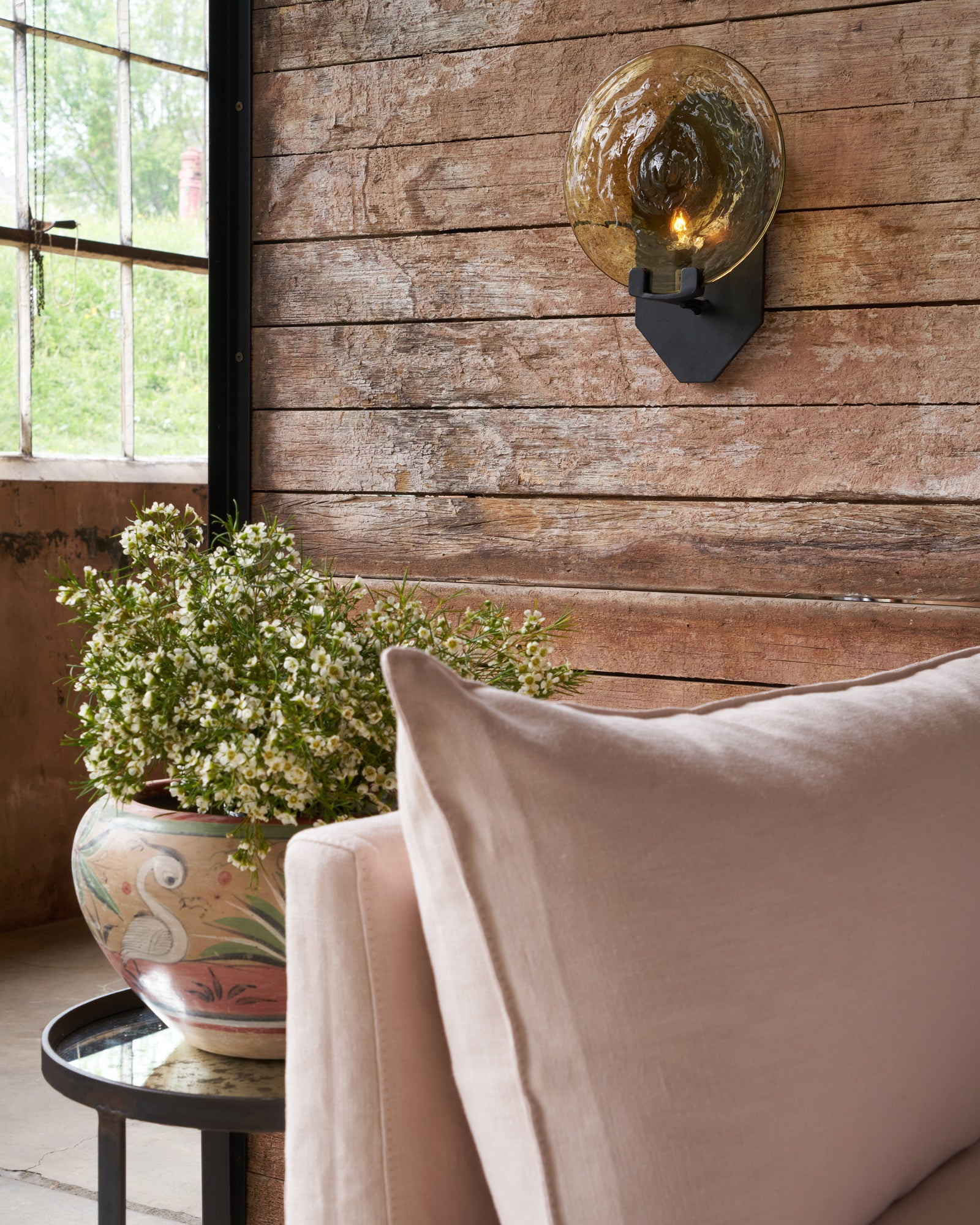  Emma Sconce on a wood wall in front of a pink sofa and a pot with white flowers.  