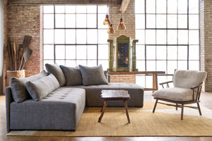  Sectional in a showroom with a yellow rug and a wood coffee table. Wood chair with an oatmeal cushion. Wood console in the background and a green wood chest. Photographed in Raven Peppercorn.  