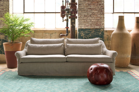A light beige upholstered loveseat sofa centered in a rustic industrial loft setting with brick columns, large windows, potted plants, and a dark red leather ottoman on a teal patterned rug. Photographed in Ashville Oatmeal.