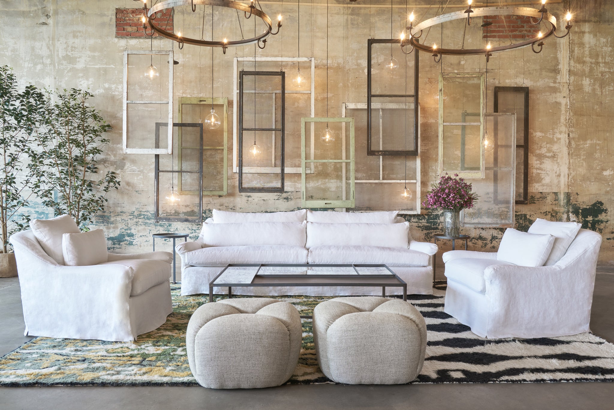  White sofa and two armchairs in front of a concrete wall with windows hanging. 2 poufs are in front of the coffee table. Photographed in Salerno Olive. 