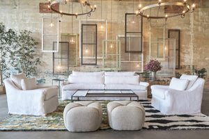  White sofa and two armchairs in front of a concrete wall with windows hanging. 2 poufs are in front of the coffee table. Photographed in Brevard Ivory. 