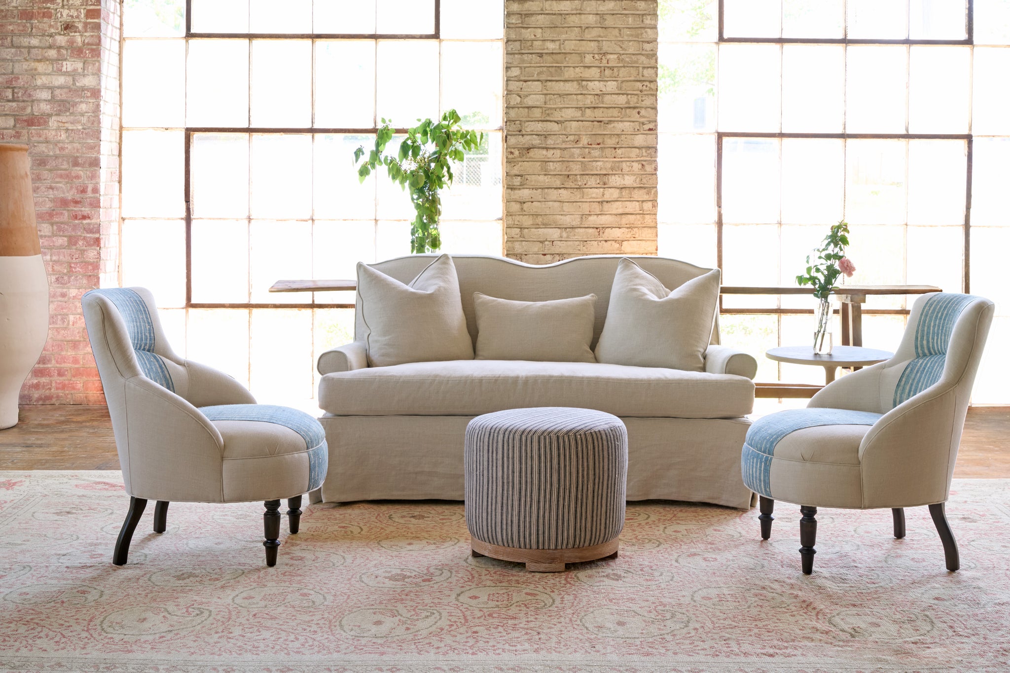  Slipcovered sofa with 3 pillows. Side table with flowers. Bench in the back, in front of 2 large windows. Two small chairs with a blue stripe in the center and a striped ottoman in front. Photographed in Noah Bone. 