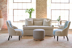  Slipcovered sofa with 3 pillows. Side table with flowers. Bench in the back, in front of 2 large windows. Two small chairs with a blue stripe in the center and a striped ottoman in front. Photographed in Noah Bone. 