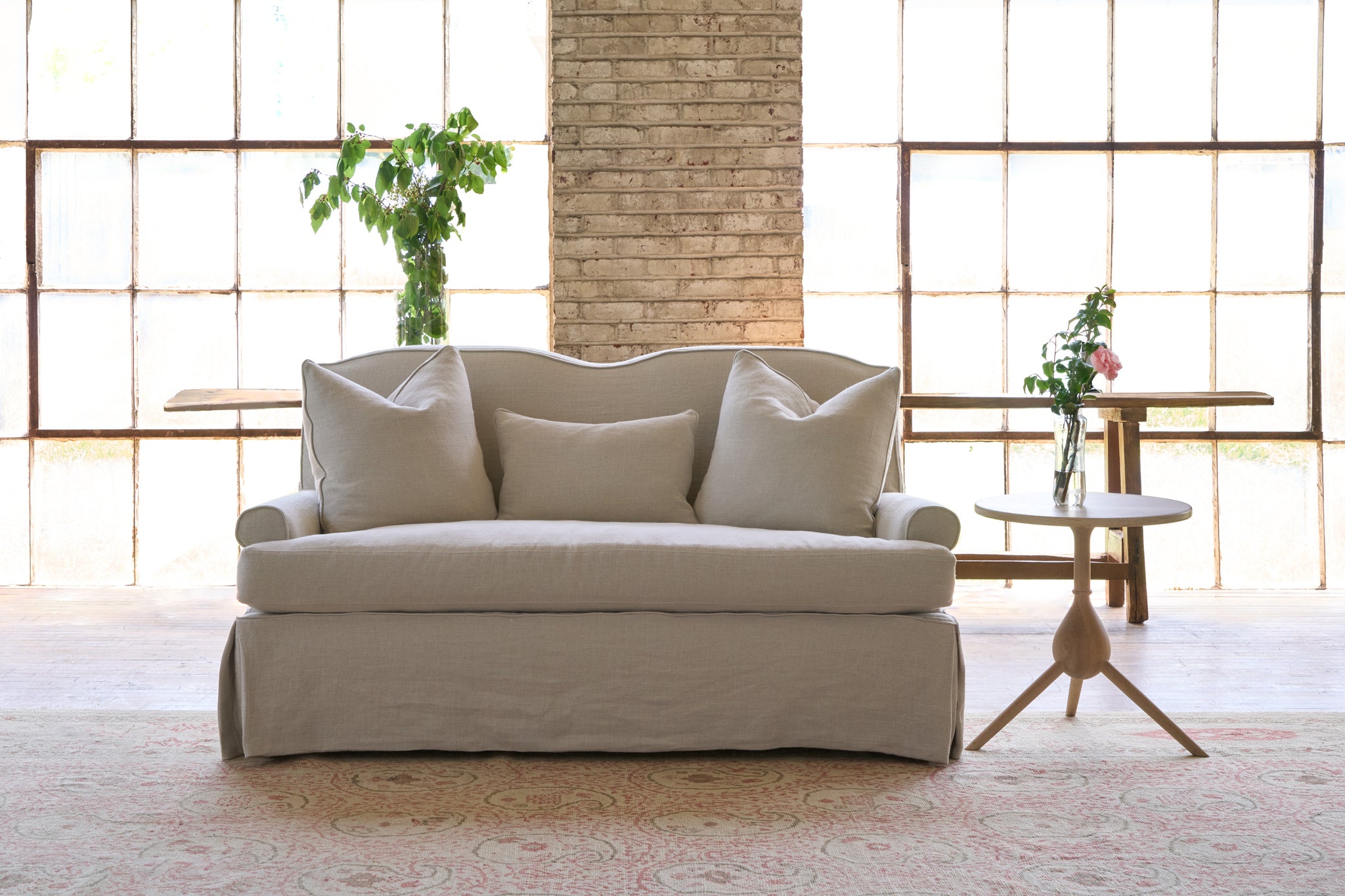  Slipcovered sofa with 3 pillows. Side table with flowers. Bench in the back, in front of 2 large windows. Photographed in Noah Bone. 