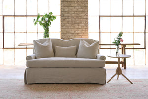  Slipcovered sofa with 3 pillows. Side table with flowers. Bench in the back, in front of 2 large windows. Photographed in Noah Bone. 