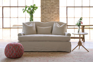  Slipcovered sofa with 3 pillows. Side table with flowers. Bench in the back, in front of 2 large windows and with a red pouf ottoman on the left. Photographed in Noah Bone. 