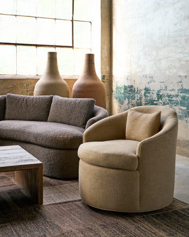 A round upholstered accent chair in a warm beige fabric sits in a styled living room next to a textured gray sofa, with two large ceramic vases and a rustic window in the background. Photographed in Harris Wheat.