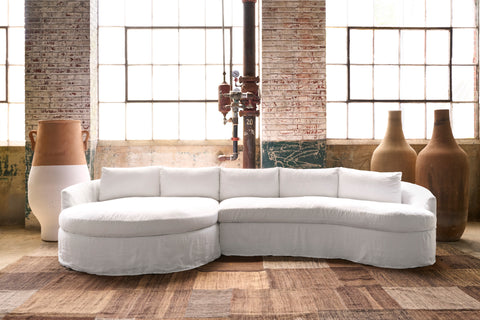 A white slipcovered curved sectional sofa in a rustic industrial loft setting with brick columns, large windows, and decorative ceramic vases. Photographed in Logan White.