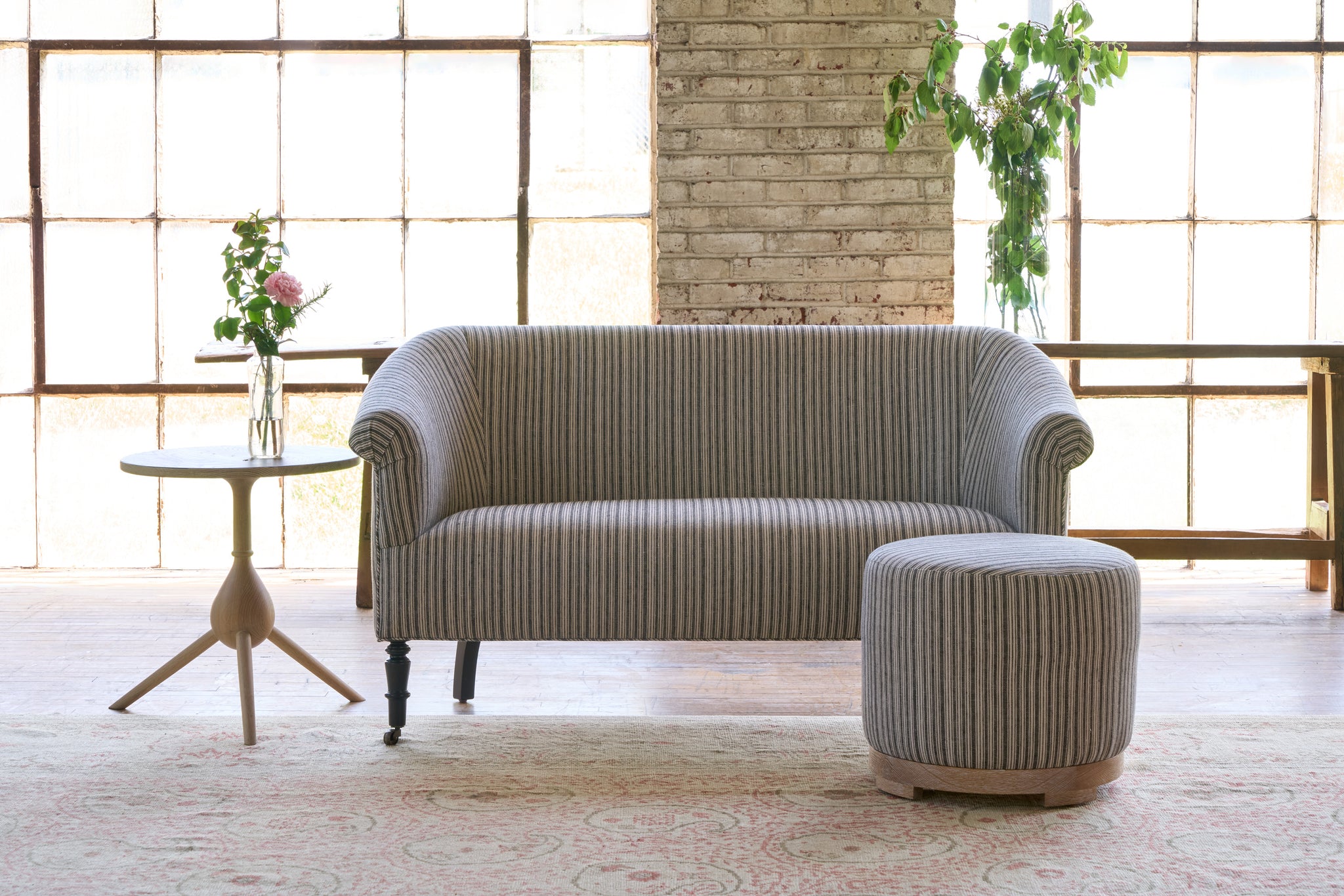  Striped sofa with a striped ottoman with a side table on the left with flowers on top. Photographed in Hickory Noire. 