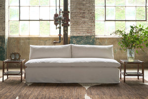  Armless sofa in cream colored fabric in front of large windows. 2 side tables with bouquet on the right and vintage double frame on the left. Photographed in Chevron Cream. 