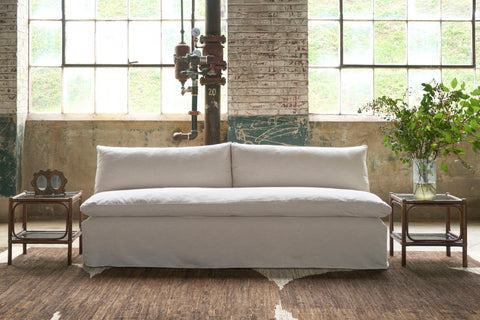 Armless sofa in cream colored fabric in front of large windows. 2 side tables with bouquet on the right and vintage double frame on the left. Photographed in Chevron Cream.