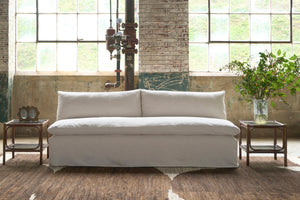  Armless sofa in cream colored fabric in front of large windows. 2 side tables with bouquet on the right and vintage double frame on the left. Photographed in Chevron Cream. 