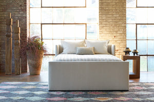  Bed in front of a large window with plant in a pot and wood sculptures on the left. Wood side table on the right. Photographed in Adler Sand. 