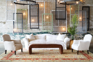  White slipcovered sofa with a brown leather bench in front. A white chair on each side. In the background, vintage window panes are hanging with lights. Photographed in Luna White. 