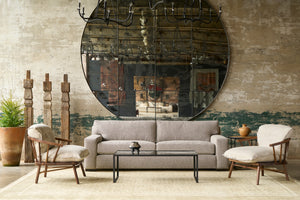  Grey sofa in front of a large mirror with 2 wood framed chairs on each side. 3 wood sculptures on the left with a plant. Photographed in Devon Oatmeal.. 