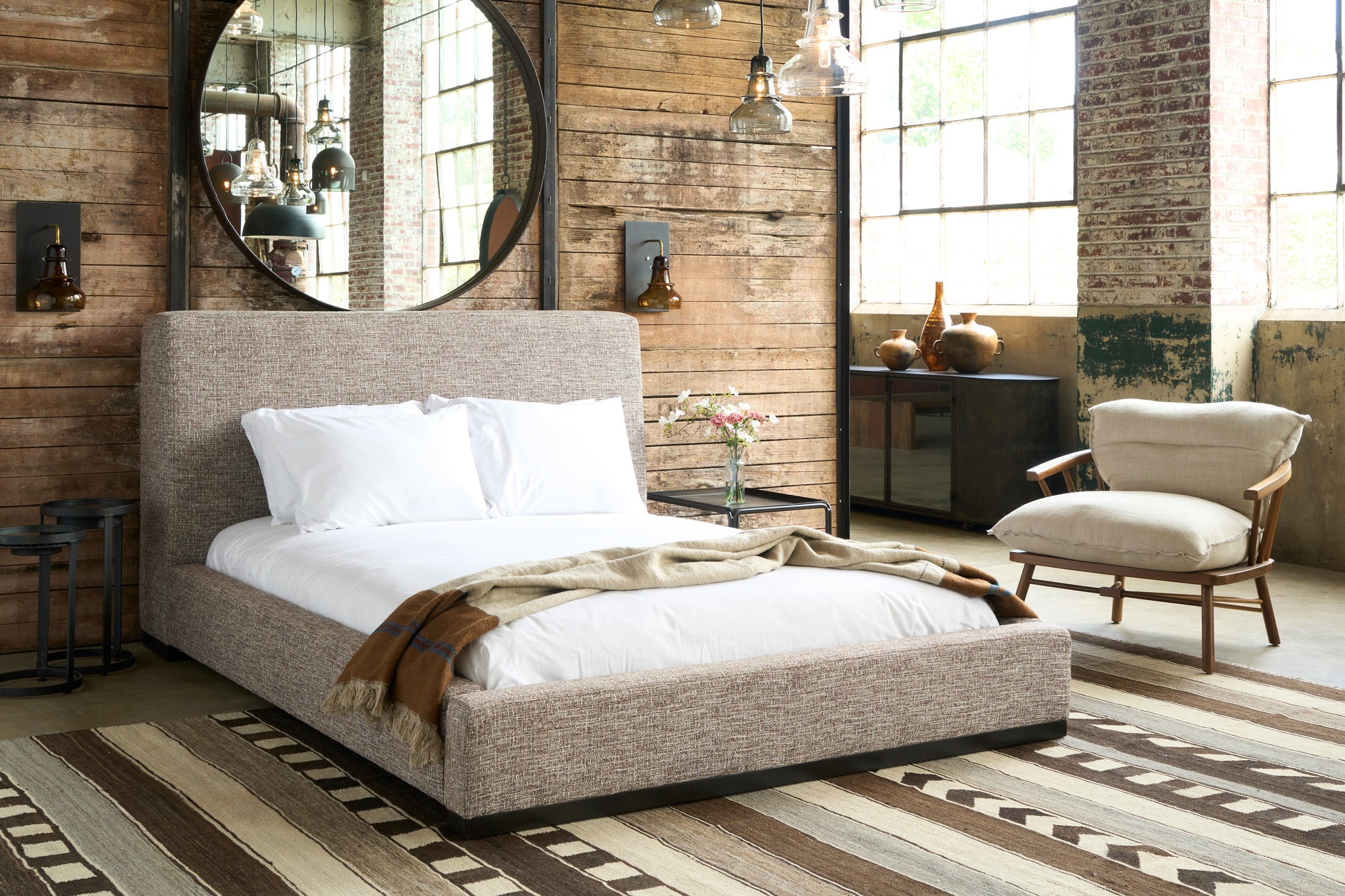  Modern bed in front of a wood wall with a squre metal table on the right with flowers on top. Sconces on the wall and a large round mirror. Wood framed chair on the right. Photographed in Verona Coffee. 