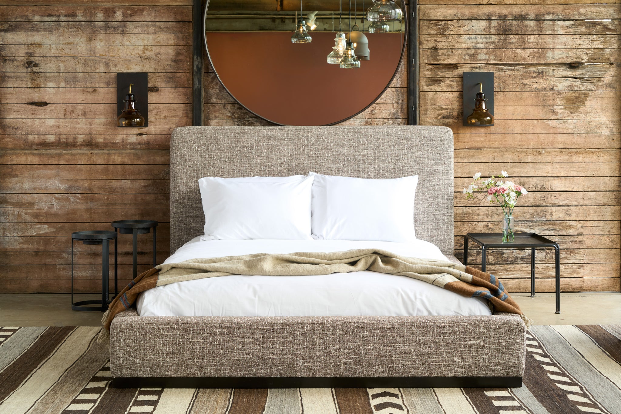  Modern bed in front of a wood wall with a squre metal table on the right with flowers on top. Sconces on the wall and a large round mirror. Photographed in Verona Coffee. 