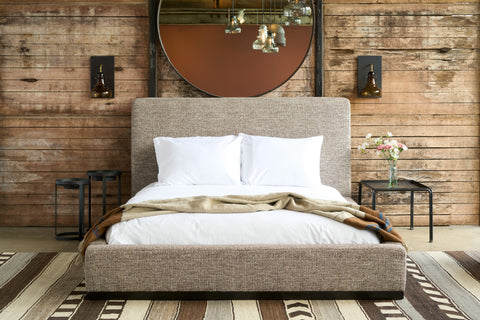 Modern bed in front of a wood wall with a squre metal table on the right with flowers on top. Sconces on the wall and a large round mirror. Photographed in Verona Coffee.