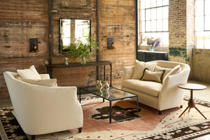  Two white loveseats facing in a showroom in front of a wood wall. Coffee table with mirrored top with 2 pots on top. Photographed in Lumi Bone. 