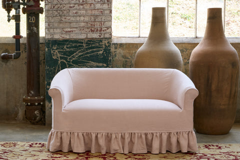 Pink slipcovered loveseat in a room with large vases and a textured wall. Photographed in Brevard Rose.