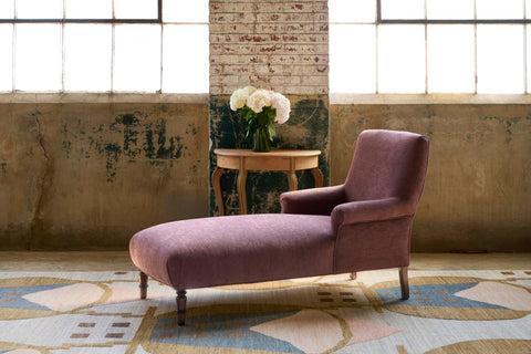 Chaise lounge in purple in a room with 2 large windows and a wood table and white flowers on top, and a multi colored rug. Photographed in Pascale Vintage Plum.