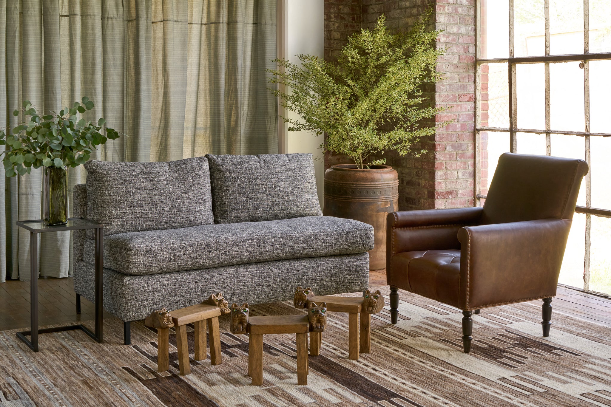  Armless sofa on a brown rug  with a brown leather chair on the right. Tree in a large pot in the back corner. 3 wooden stools with dog heads in front . Photographed in Verona Black. 