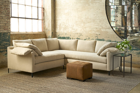 Beige sectional sofa with a brown ottoman in a room with large windows and exposed brick walls. Photographed in evon Oatmeal.