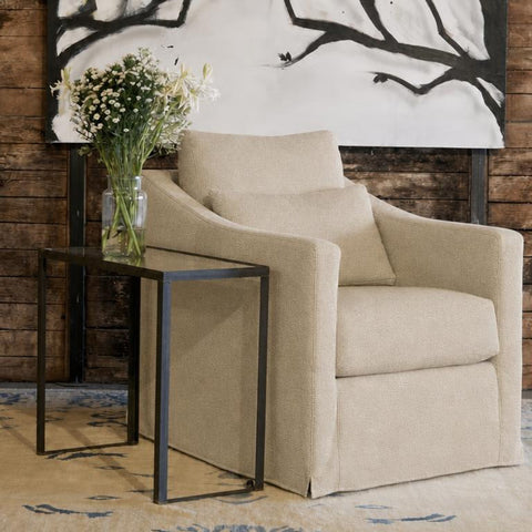 A beige upholstered armchair with a matching back cushion, styled in a living room setting next to a black metal side table with a vase of flowers, set against a rustic wood plank wall and a large black-and-white abstract painting.