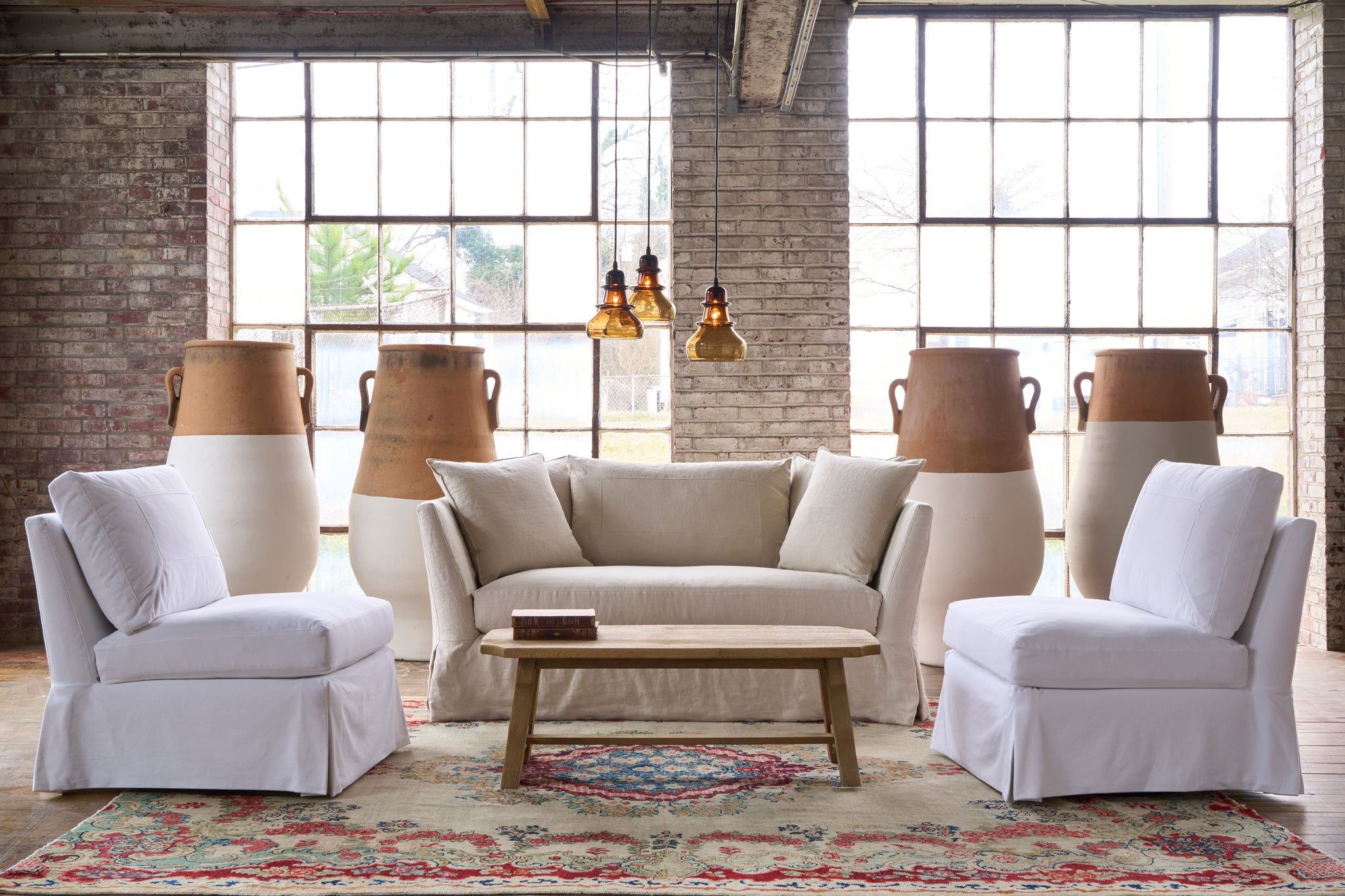  Loveseat in front of a wall and windows with large painted pots. 3 brown glass pendants are hanging. Two armless chairs in denim on each side. Photographed in Noah Bone. 