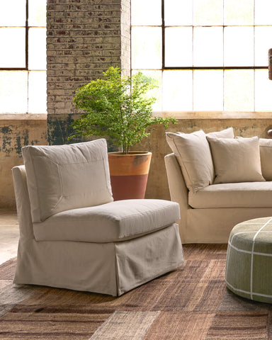 A beige slipcovered armchair with a skirted base is shown in a styled living room setting with a matching sofa, a potted plant, and a patterned rug. Photographed in Denim Natural.