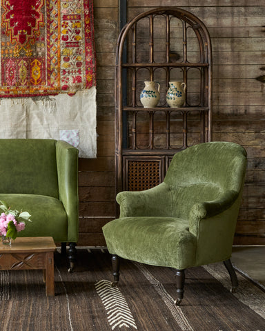 Green leather loveseat with a green velvet chair in front of a wood wall with some rug art. Wood coffee table with a drawer in front. Rattan cabinet on the right. Photographed in Velluto Olive.