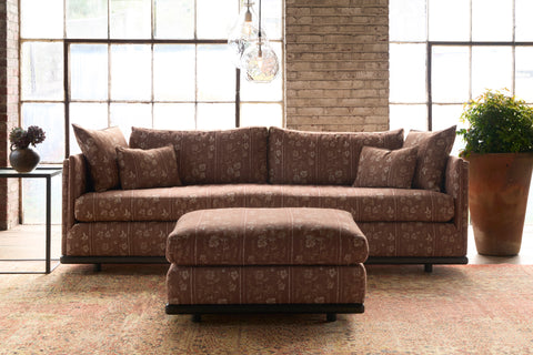 Brown patterned sectional sofa with matching ottoman in a room with large windows and a stone wall. Photographed in Foret Rust.