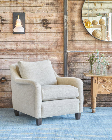 Oatmeal colored chair in front of a wood wall with a white sconce light behind and a round mirror on the right. Photographed in Bellamy Oatmeal.