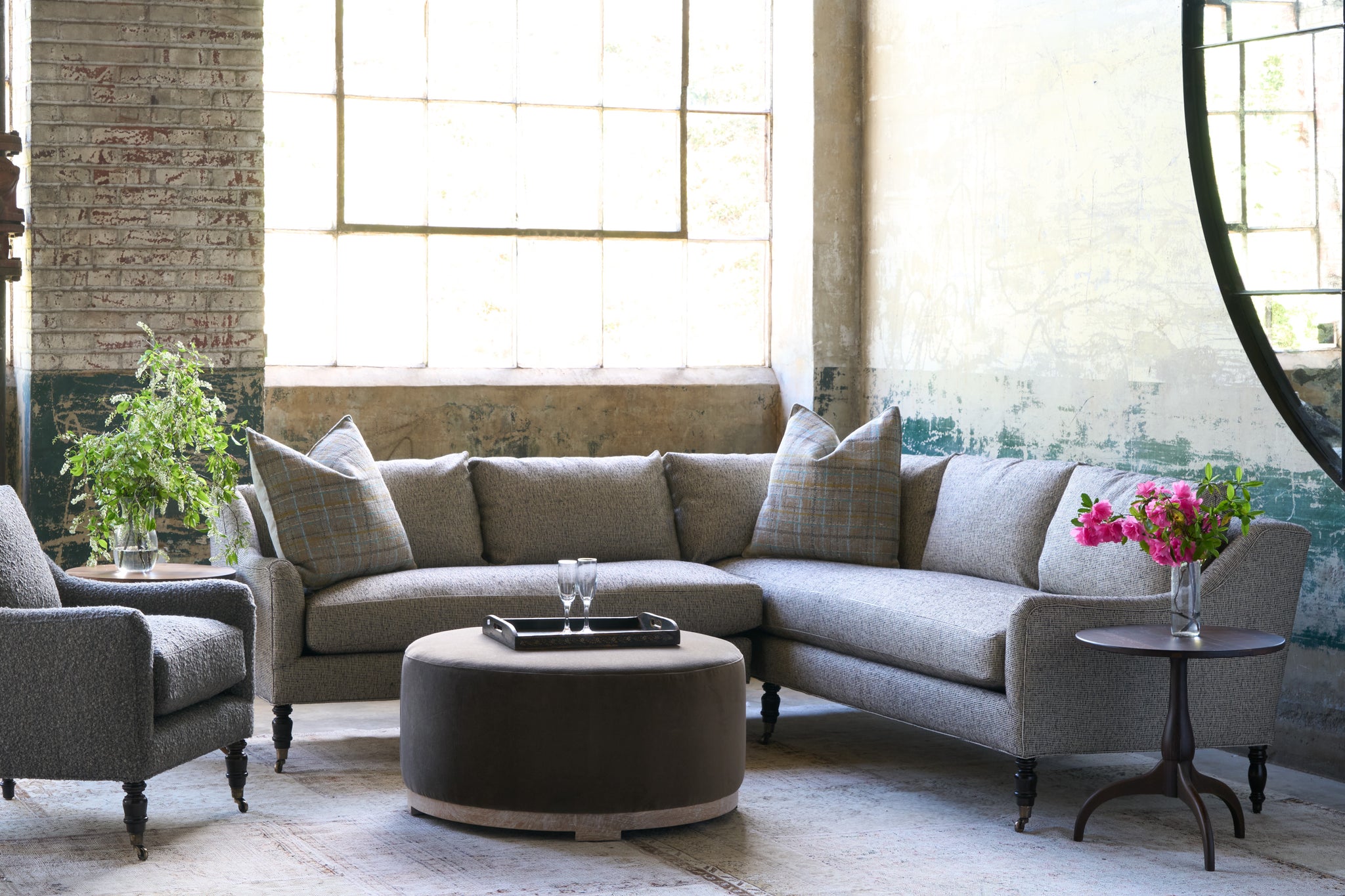  Sectional sofa in front of a large window with an ottoman in front. Tray on top with 2 glasses. Chair on the left and wood side table on the right with pink flowers on top. Photographed in Ferrara Charcoal. 