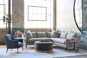  Sectional sofa in front of a large window with an ottoman in front. Tray on top with 2 glasses. Chair on the left and wood side table on the right with pink flowers on top. Photographed in Ferrara Charcoal. 