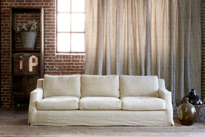  The Hayden slipcovered sofa in Lan Oatmeal is in a room with a brick wall and a grey curtain covering the windows. There is a bookcase to the left with some flowers and signs, to the right there are some vintage glass vessels.  Photographed in Lan Oatmeal. 