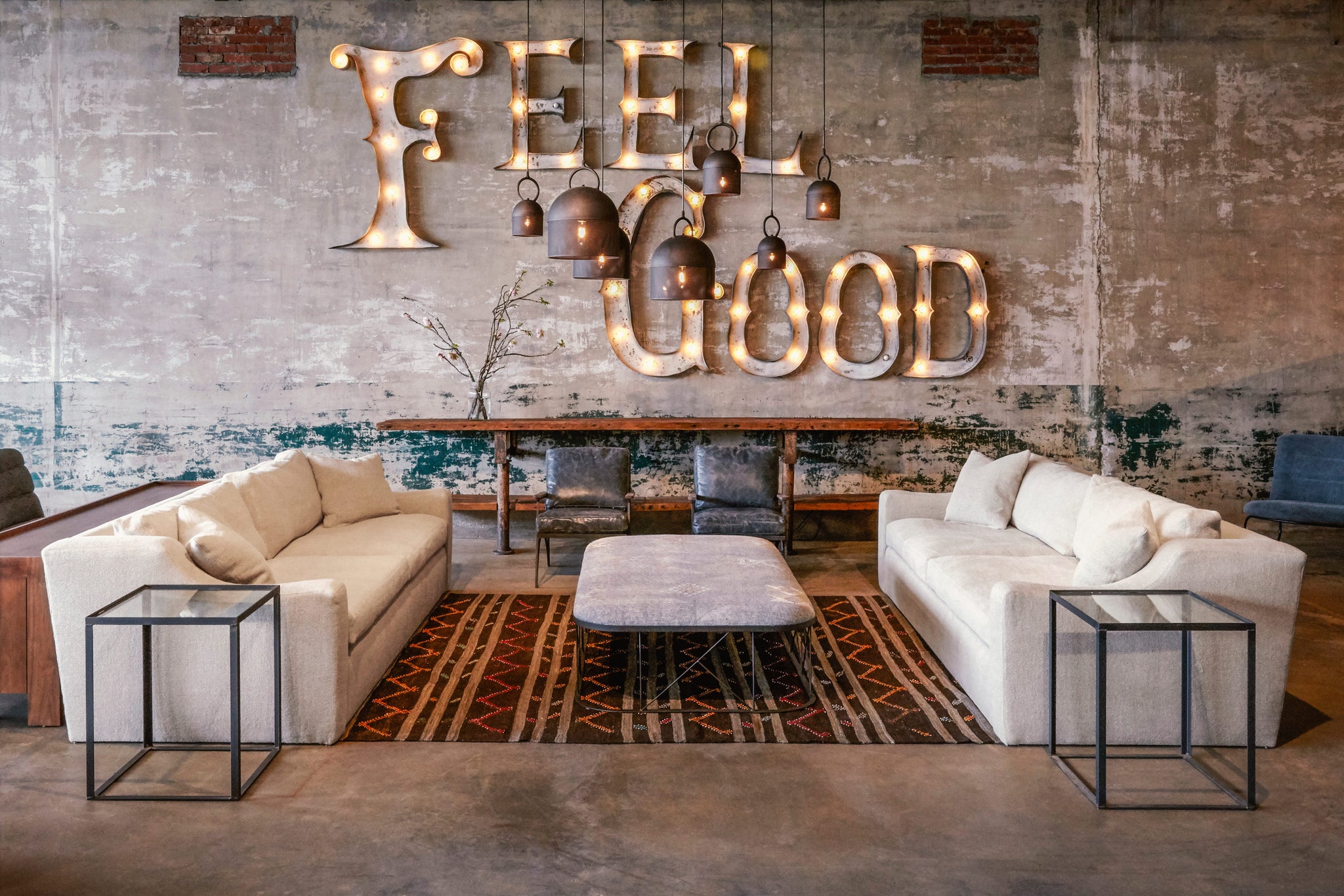  Two Grant sofas Brevard Burlap are facing each other in a large showroom space. There is a Feel Goof metal sign with lights hanging on the back concrete wall. A large rectangular ottoman is in the middle and two square side tables at the end of each sofa. There are 2 leather chairs in front of the wall. Photographed in Lan Oatmeal. 