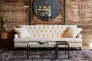  The Henderson sofa is in Avery Oatmeal in front of a wood wall with a round mirror hanging. The coffee table in front is a 2 piece Welders coffee table in glass and metal. Photographed in Avery Oatmeal. 
