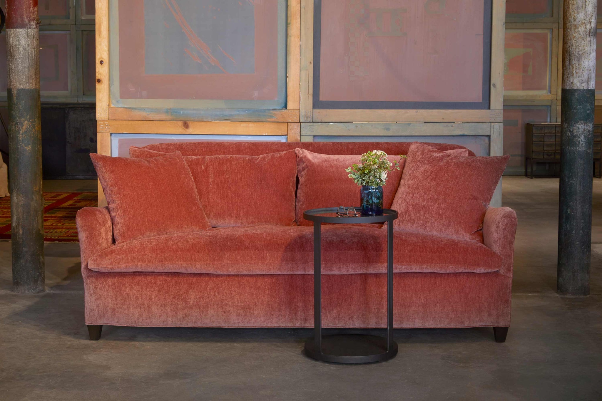  The Hazel loveseat is upholstered in Lacey French Rose with a cantilever side table in front. There is a vase with flowers and glasses on the table. The background is a wall with vintage pink sunscreens.  Photographed in Lacey French Rose. 
