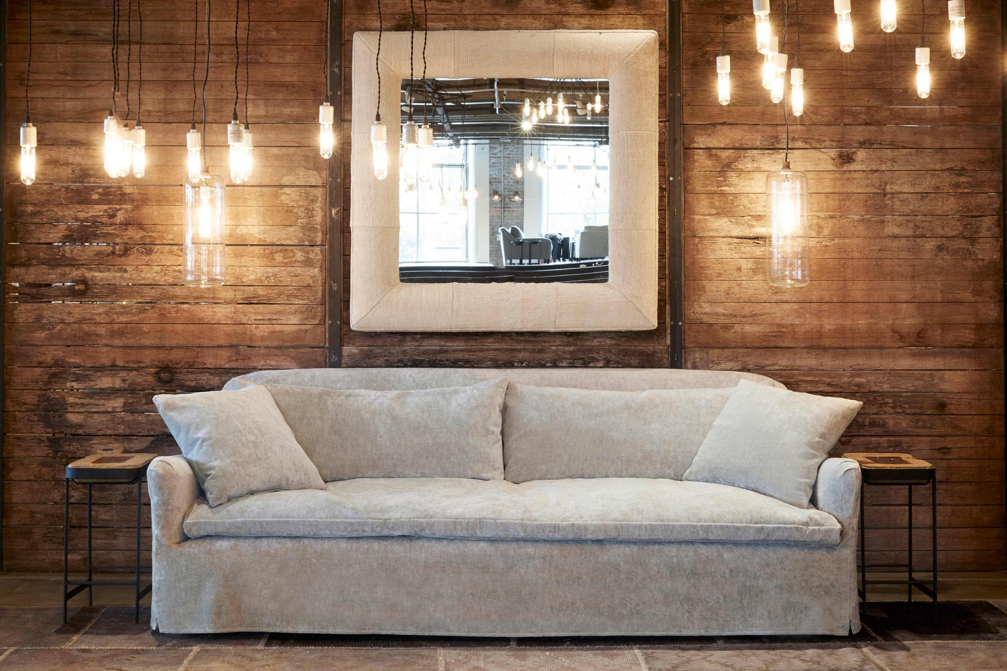  The Hazel sofa is slipcovered in Velluto Stone in front of a wood wall with a square mirror with a fabric frame. There are lots of small lights hanging and 2 Cylinder lamps on each side of the sofa. Photographed in Velluto Stone. 