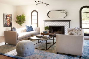  Living room, daytime lighting with 2 facing Kenso sofas in Mariet Natural. A fireplace with an oval Cooper mirror hanging above. Large rectangular coffee table with books on top. A Pouf Ottoman in grey fabric in front. A painting on the back wall and a tree in the corner. Photographed in Mariet Natural. 