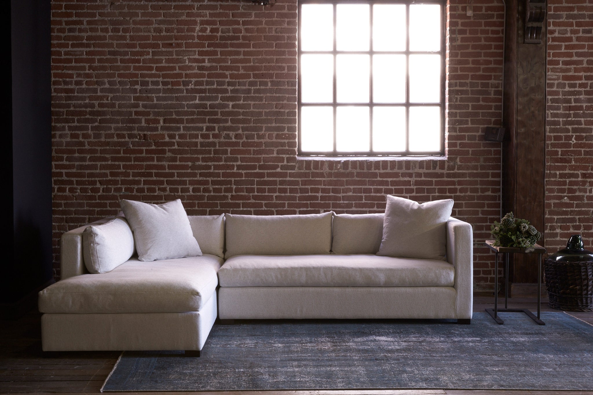 The Agosto sectional, left arm facing, in Adler Sand is in a room with dim light, in front of a brick wall with a large window behind. Photographed in Adler Sand. 