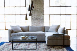  This a light grey Agosto 2pc sectional right arm facing  on top of a  floral rug with a blue trim in front of two big large window during daylight, staged with coffee table in front of sectional and side table in front of the bumper sectional and a Chandelier hanging above the coffee table. Photographed in Adler Sand. 