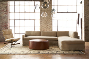  Allister Sectional in a large room with 2 big windows. There is a metal chair with a cream cushion on the left and a brown ottoman in the center. Photographed in Brevard Burlap. 